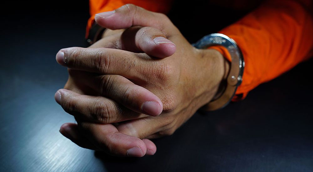 Clasped hands with handcuffs rest on a table, highlighted by a focused light in a jail cell.