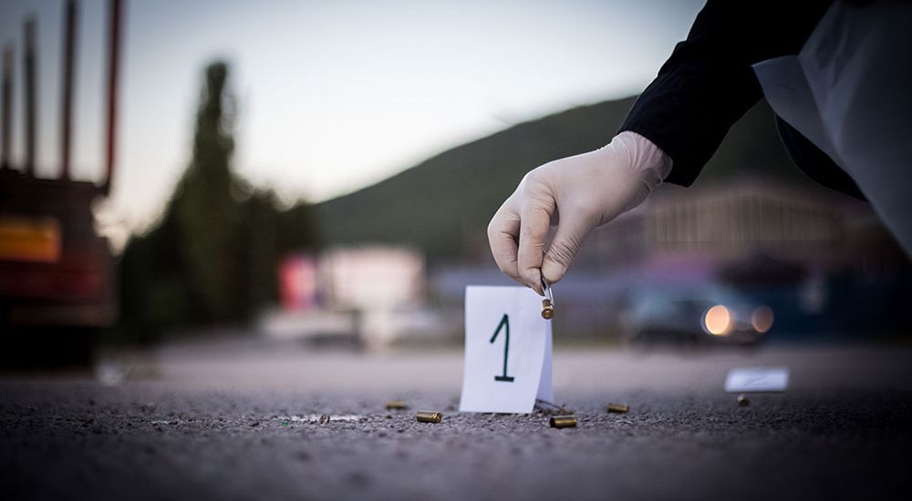 A gloved hand places a bullet casing next to a numbered evidence marker on a road, with blurred vehicles and trees in the background, suggesting a crime scene investigation.