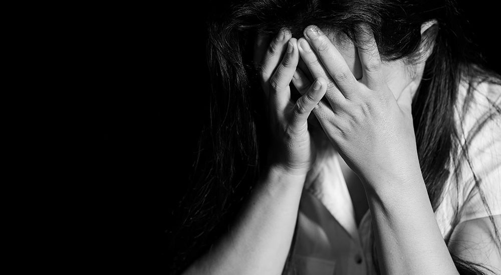 A person with long hair covers their face with both hands, conveying distress. They are set against a stark black background, highlighted by dramatic lighting on one side.
