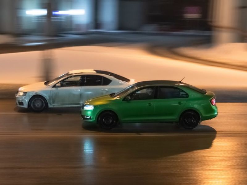 Two cars in motion on city street at night. Racing at high speed on public road. White and green sedans drive on wet winter road