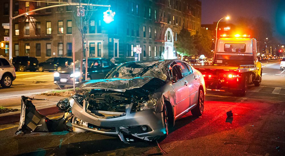 A damaged car sits in the middle of a street at night, with parts strewn around and a tow truck positioned behind it, indicating a recent accident in an urban area.