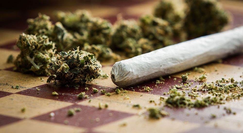 A hand-rolled cigarette rests beside dried cannabis buds on a checkered surface, suggesting preparation for smoking.