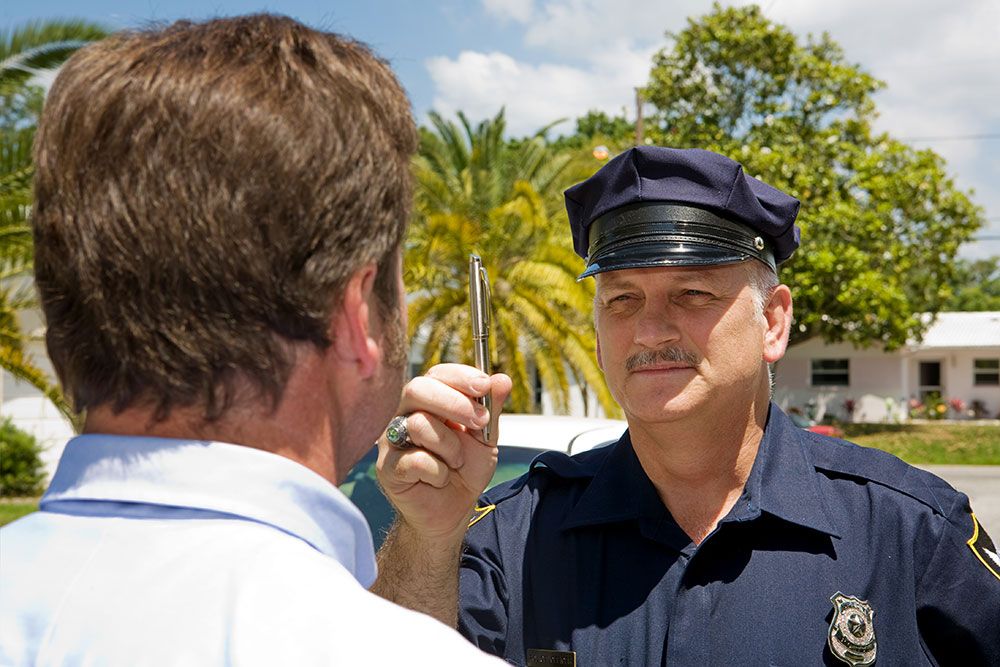A police officer is performing a sobriety test on a man by holding out a pen, against a suburban backdrop with palm trees.