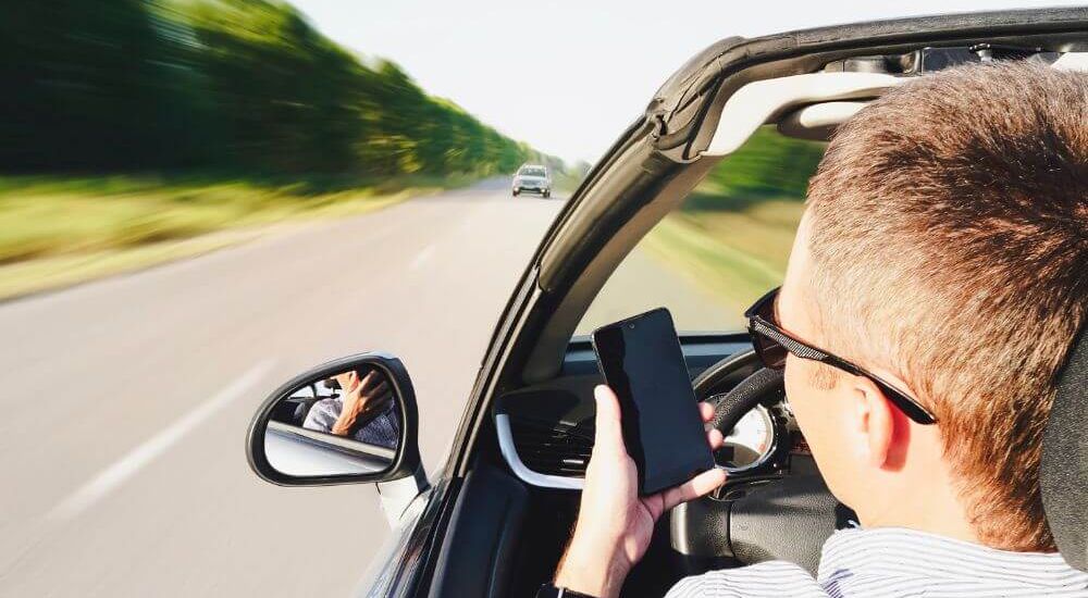 A man driving a car, it seems to be speeding while holding a phone with one hand and not paying attention to the road.