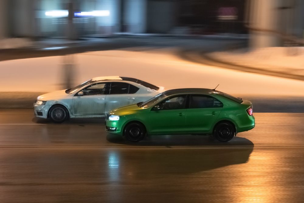 Two cars in motion on city street at night. Racing at high speed on public road. White and green sedans drive on wet winter road