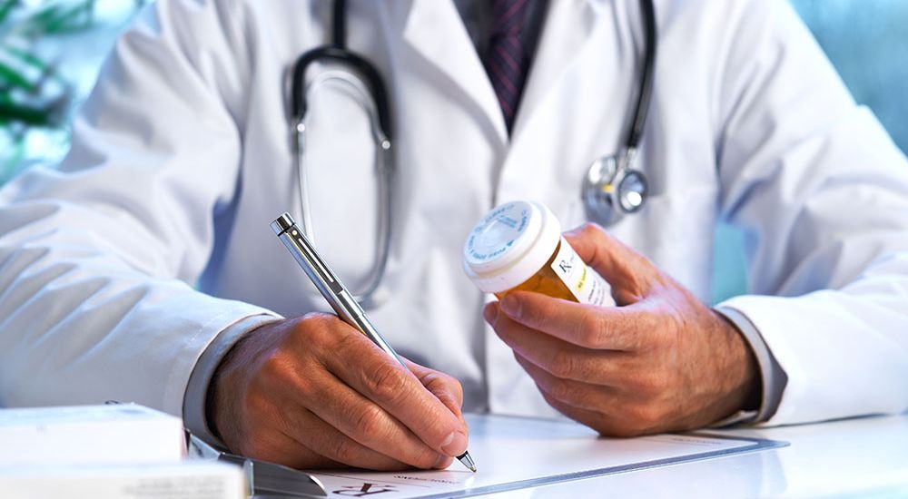 A doctor is writing on a prescription pad and holding a medication bottle, in a well-lit office with medical equipment in the background.
