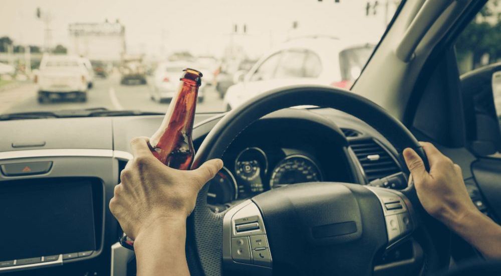 A person holds a brown bottle while driving a car on a busy road, surrounded by other vehicles and traffic lights.