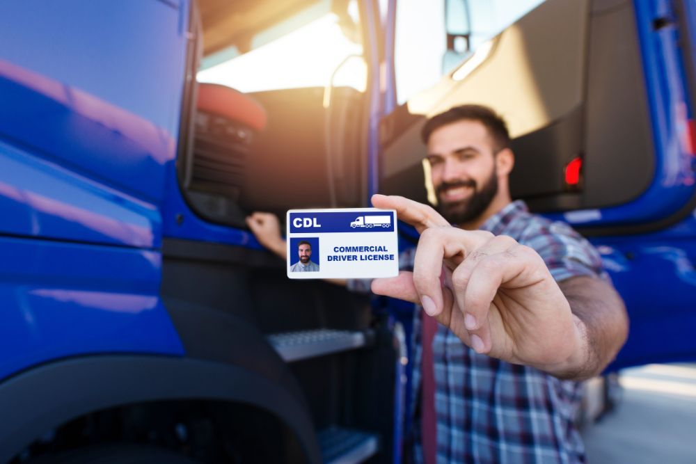 A smiling man displays his Commercial Driver License (CDL) outside a blue truck, highlighting his qualification to operate commercial vehicles.