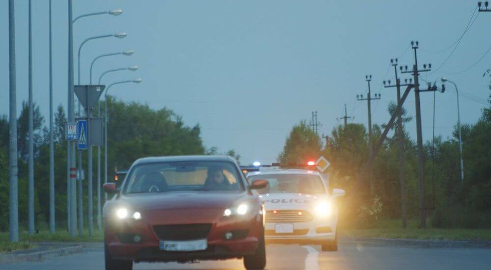 A red car is driving forward on a road, while a police car with flashing lights approaches from behind. The setting is suburban, with streetlights and trees lining the sides.