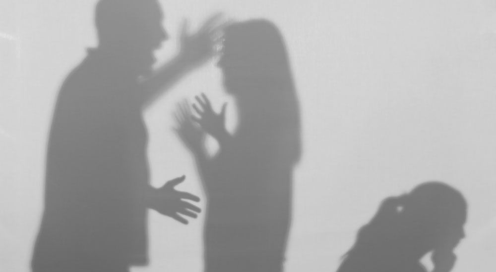 Silhouetted figures appear to be engaging in a heated argument, with the man gesturing toward the woman, against a plain backdrop; a third figure's shadow is also visible.