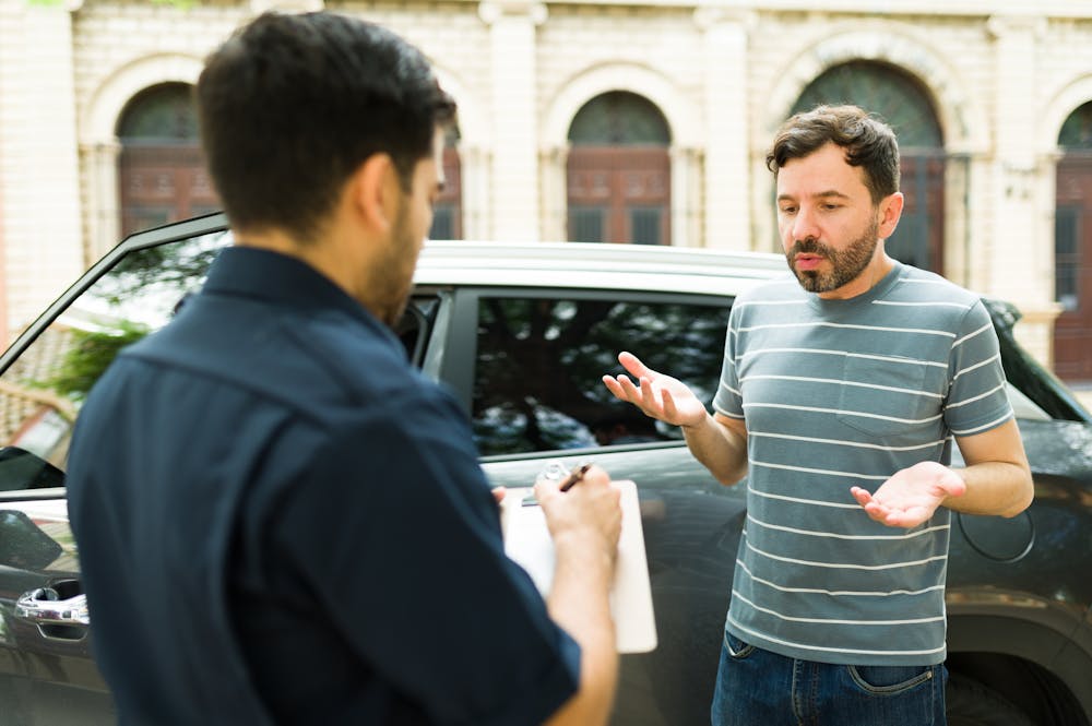 A man in a striped shirt gestures with his hands while talking to another man taking notes. They are next to a parked car outdoors, surrounded by buildings.