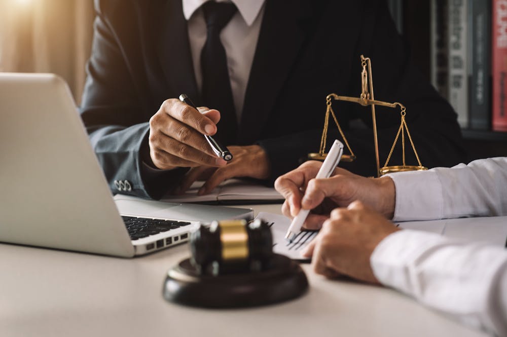 Two individuals in formal attire, one writing with a pen and the other holding a pen, are engaged in discussion at a desk featuring a laptop and a wooden gavel. A balance scale is also present.