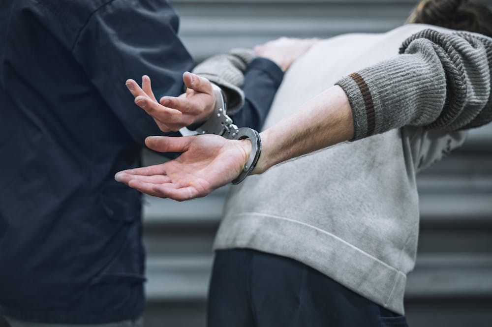 A person with their hands cuffed behind their back is being restrained by another individual. The setting appears to be an urban environment with a gray background.